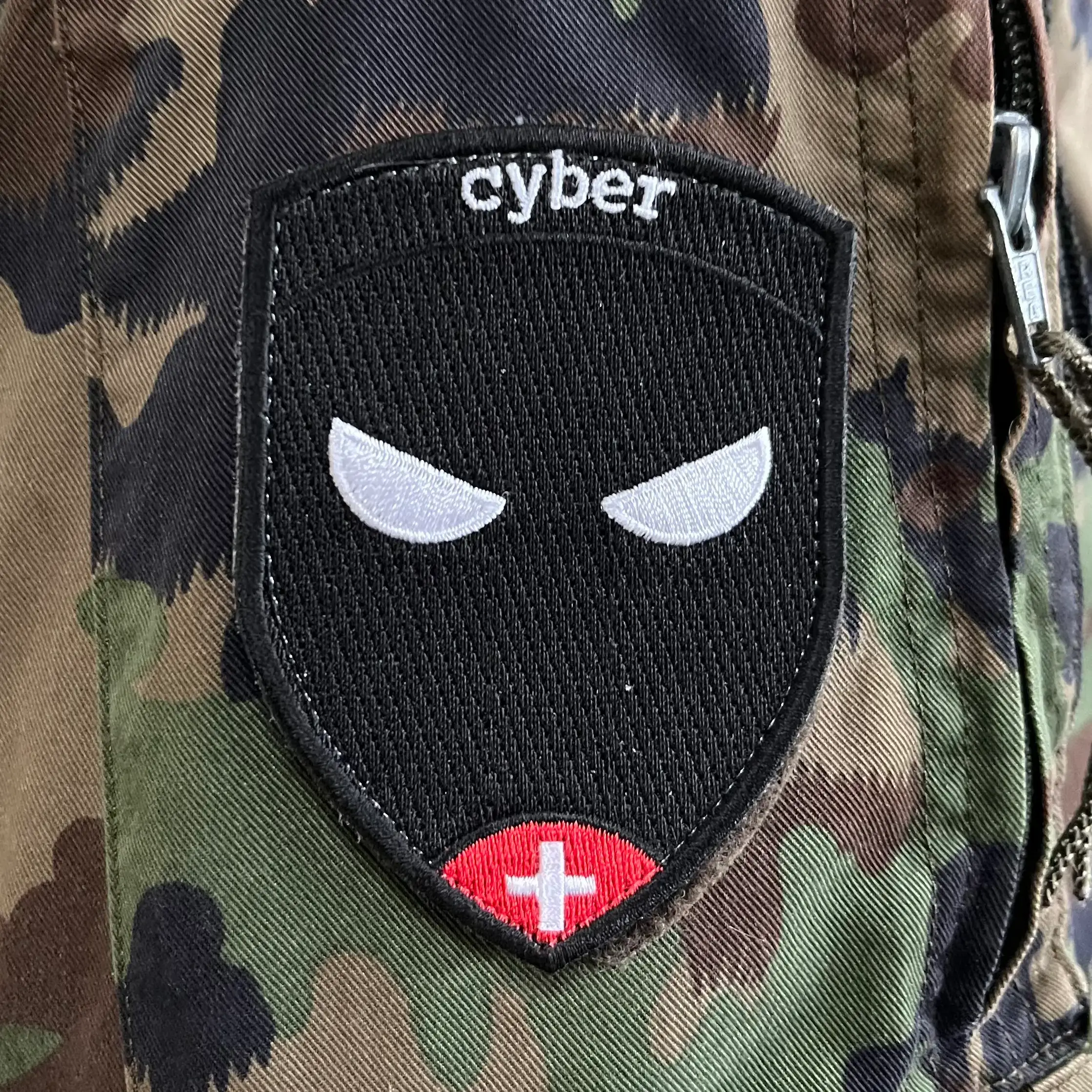Militärisches Abzeichen von RS, gekauft und hergestellt auf RS Badge. Das Abzeichen ist einfach und schlicht, auf einem schwarzen Hintergrund steht Cyber. Unten auf dem Abzeichen sehen wir ein Schweizer Kreuz auf einem roten Arroundi und in der Mitte des Abzeichens sind zwei offene Ninja-Augen abgebildet.